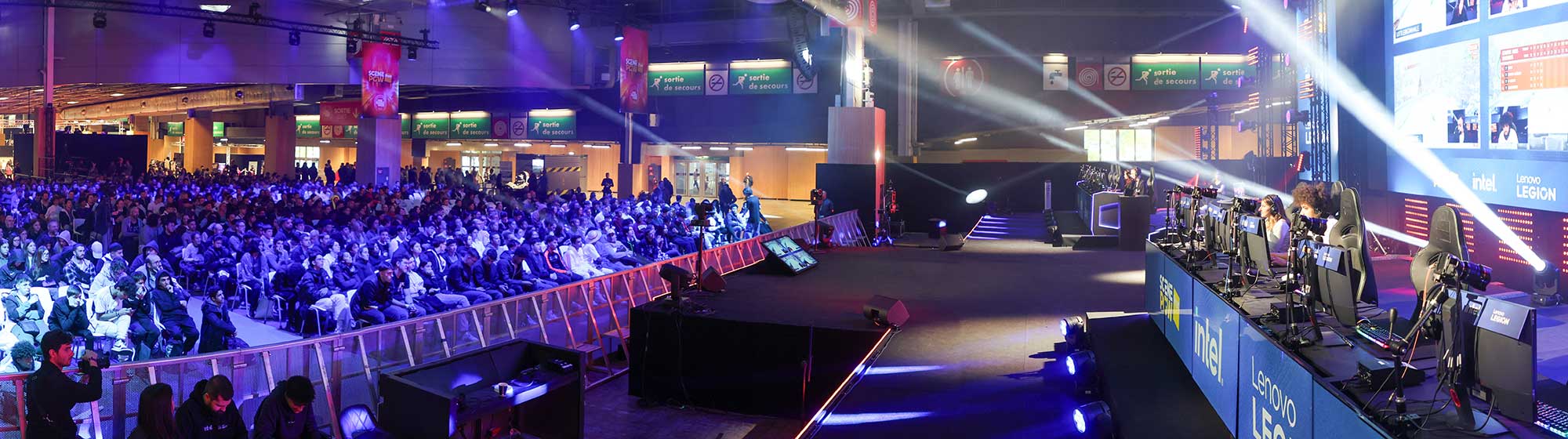 A seated crowd watches gamers play video games on a stage with a giant screen