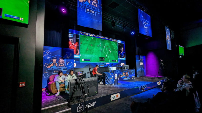 des jeunes jouant un match en ligne sur un jeu vidéo de football dans une salle où le match est rejoué sur un écran géant