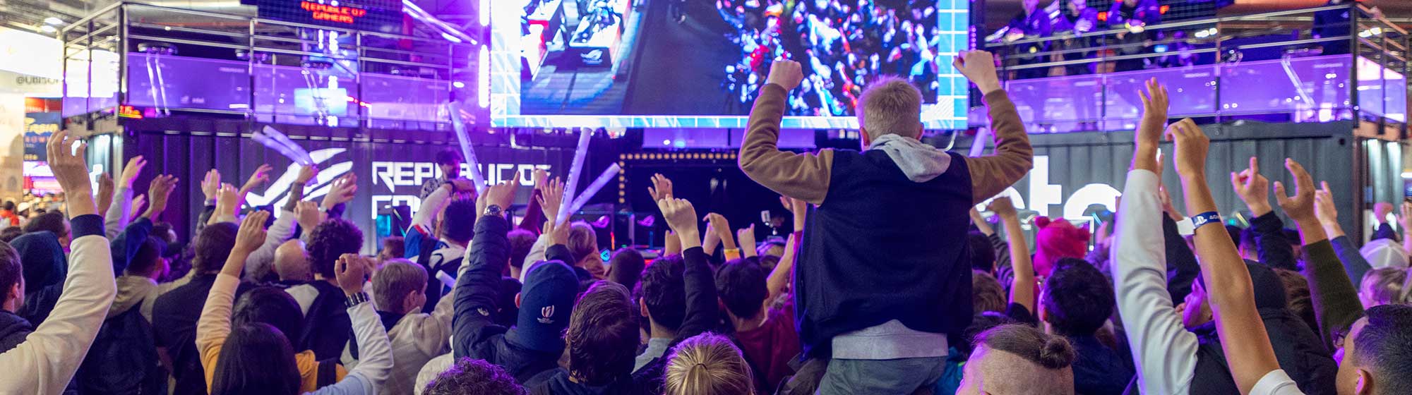 A parent carries his son on his shoulders amidst a cheering crowd outside a stand at Paris Games Week.