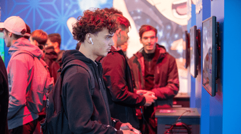 Jeune concentré debout jouant à un jeu vidéo avec des gens en arrière-plan